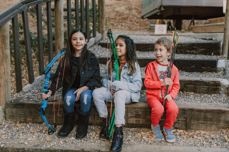 Three children sitting on steps holding bows and arrows, enjoying archery practice in a forest setting.